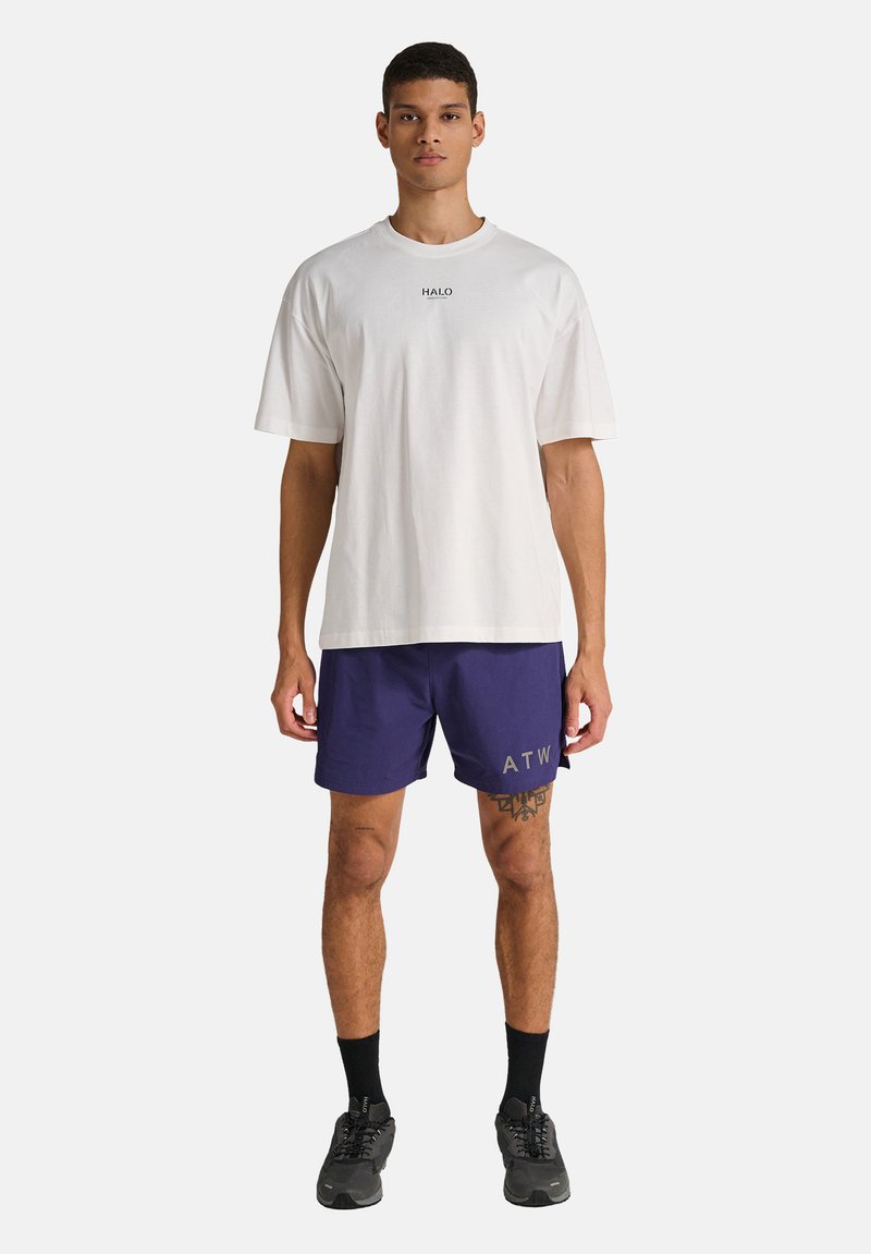 Mann steht, trägt ein weißes HALO-T-Shirt, marineblaue Shorts mit ATW-Schriftzug, schwarze Socken und schwarze Turnschuhe vor einem schlichten weißen Hintergrund.