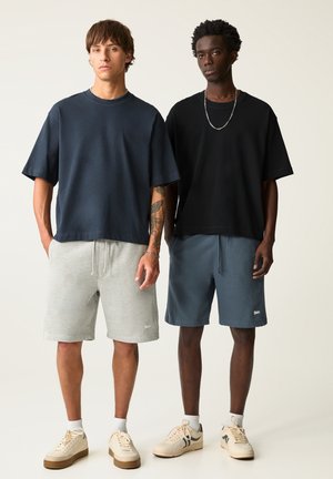 Zwei Männer stehen nebeneinander und tragen lässige Outfits. Der eine trägt ein dunkelblaues T-Shirt und graue Shorts; der andere trägt ein schwarzes T-Shirt und blaue Shorts.