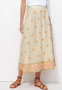 Jupe beige avec broderies florales orange, ornée d'un ourlet à motifs colorés. Accompagnée de sandales à talons bruns.
