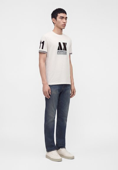 Мъж стои, носейки бяла тениска Armani Exchange с черно лого и номер 11 на ръкава, сини дънки и бели маратонки.