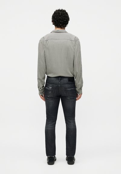 Άνδρας με σγουρά μαλλιά, ντυμένος με ένα μακρυμάνικο πουκάμισο σε ασπρόμαυρο καρό, σκουρόχρωμα φθαρμένα τζιν και μαύρα παπούτσια, στέκεται γυρισμένος από την πλάτη.