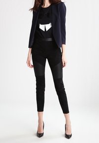 Mujer con pantalones ajustados negros con paneles de rodilla acanalados, camiseta gráfica negra, bléiser azul marino y tacones altos negros, de pie contra un fondo blanco.