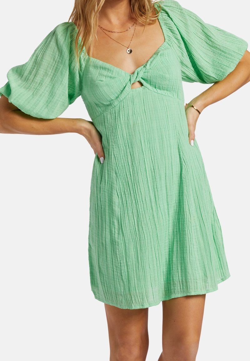 Robe vert pâle avec manches bouffantes, tissu texturé et un détail nœud au niveau de la poitrine. Silhouette évasée en ligne A, longueur au genou.