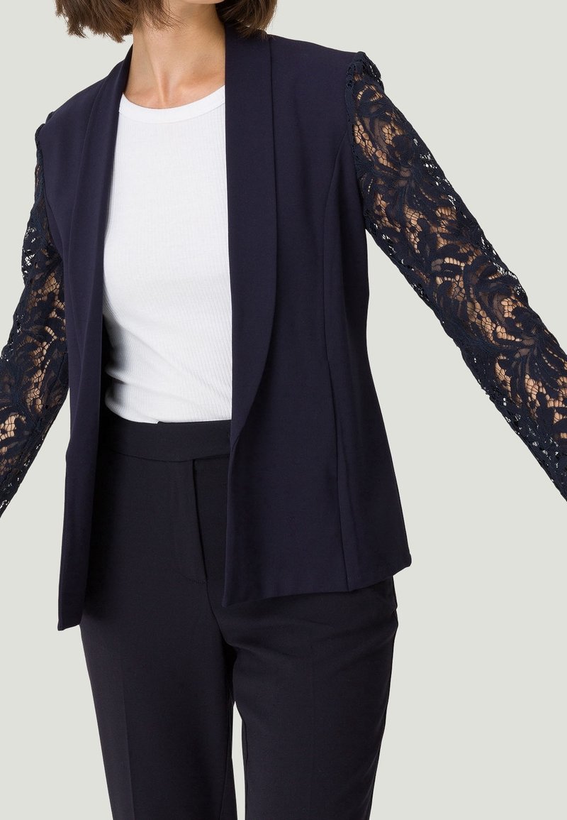 Blazer azul marino con mangas largas de encaje, que presenta un diseño entallado y un frente abierto, combinado con una parte superior blanca ajustada debajo.