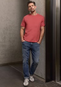 Homme en t-shirt corail et jean bleu appuyé contre un mur en béton près d'un ascenseur, les mains dans les poches.