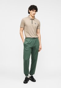 Μπεζ πόλο μπλουζάκι με φερμουάρ και λογότυπο, σε συνδυασμό με σκούρο πράσινο παντελόνι φόρμας με πλαϊνές ρίγες. Μαύρα αθλητικά παπούτσια ολοκληρώνουν την εμφάνιση.