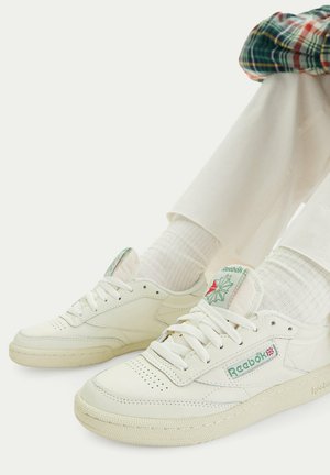 Persona con zapatillas blancas Reebok, calcetines blancos y pantalones beige sentada con las piernas cruzadas frente a un fondo liso.