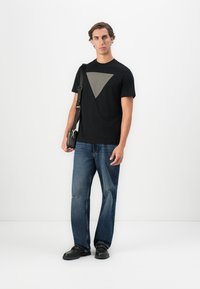 Hombre de pie que lleva una camiseta negra con un triángulo gris, jeans azules, zapatos negros y lleva una bolsa de hombro negra, sobre un fondo blanco.
