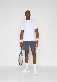 Valkoinen lyhythihainen t-paita, tummansiniset shortsit, pitää tennismailaa, valkoiset hihansuut ja urheilukengät. Yksinkertainen muotoilu, urheilullinen istuvuus.