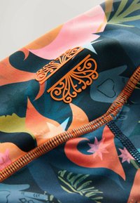 Šarena tkanina za kupanje s cvjetnim i apstraktnim uzorkom u narančastoj, ružičastoj i zelenoj boji. Uključuje detaljni narančasti simbol i kontrastne šavove.