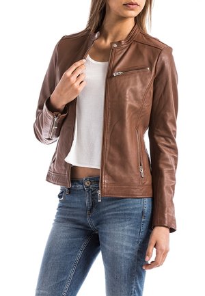 Kvinde iført en brun læderjakke over en hvid cropped skjorte og blå jeans, der holder jakkeens lynlås nær kraven.