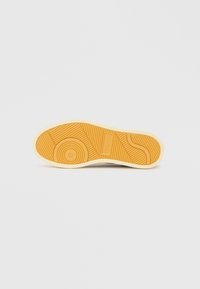 Semelle de chaussure avec une surface en caoutchouc jaune texturée, présentant des motifs circulaires et linéaires, et un bord blanc tout autour.