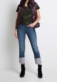 Chaleco acolchado morado oscuro sobre una camiseta verde con patrón de camuflaje, combinado con jeans azules con los puños enrollados y botas negras hasta el tobillo.