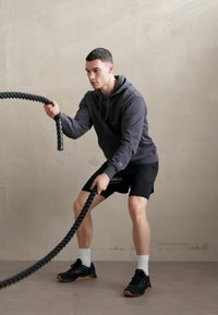 Un uomo in una felpa scura e pantaloni corti si allena con robuste corde da battaglia nere, in piedi su una superficie grigia liscia contro un muro chiaro.