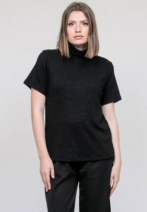 Jeune femme aux cheveux mi-longs portant un pull à col roulé noir à manches courtes et un pantalon noir, debout devant un fond uni.