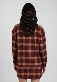 Donna con lunghi capelli mossi e scuri che indossa un abito a maniche lunghe a quadri marrone e beige, in piedi con le mani rilassate ai lati, di spalle rispetto alla fotocamera.