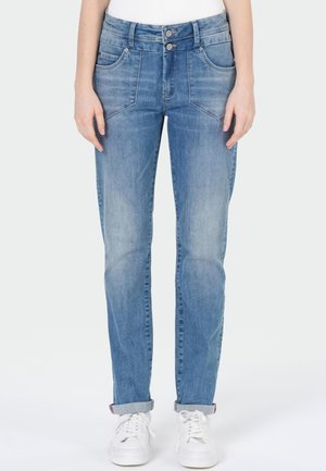 Femme portant un jean bleu clair droit avec une taille à double bouton et des baskets blanches, debout, les mains détendues le long du corps.