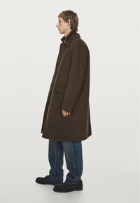 Manteau en laine marron avec un col haut, des poches latérales et une coupe légèrement surdimensionnée, associé à un jean en denim bleu et des chaussures noires.