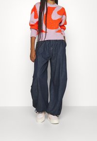 Pull en tricot violet clair avec de larges tourbillons orange, associé à un pantalon en denim foncé ample et des baskets pastel.