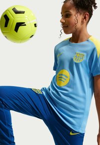 Niña con el uniforme azul y amarillo del FC Barcelona equilibrando un balón de fútbol neon amarillo y negro en su rodilla levantada.