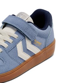 Blauwe suède sneaker met witte chevron-accenten, een marineblauwe kraag, vetersluiting en een zool van gummy rubber. Hummel-logo in gouden detail.