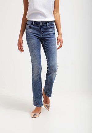 Vrouw draagt slim-fit blauwe spijkerbroek, witte mouwloze top en beige puntige platte schoenen, staand tegen een effen witte achtergrond.