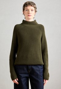 Maglione lavorato a maglia in verde oliva scuro con colletto alto e texture a costine. Le maniche lunghe presentano aperture per il pollice, abbinato a jeans scuri.
