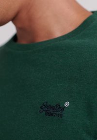 Nahaufnahme einer Person, die ein grünes SuperDry T-Shirt mit einem kleinen schwarzen Logo auf der Brust trägt.