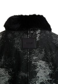 Abrigo negro con estampado que presenta un cuello de pelaje sintético y una tela texturizada. Incluye un parche rectangular de la marca en la parte posterior.