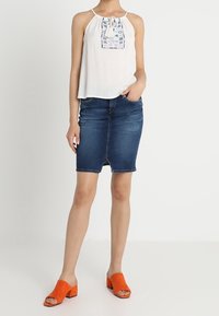 Biały top na halter z haftowanymi detalami, niebieska dżinsowa spódnica ołówkowa i pomarańczowe zamszowe mule na blokowym obcasie. Płaskie tło.