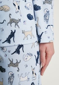 Hellblauer Stoff mit einer Vielzahl von illustrierten Katzen in verschiedenen Posen und Farben, ergänzt durch florale Akzente und einem lässigen Schnitt.