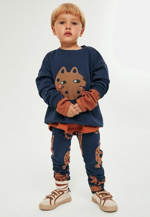 Granatowa bluza z grafiką brązowego kota, nałożona na długi, rdzawy rękaw. Granatowe spodnie z wzorami kotów. Białe sneakersy z brązowymi akcentami.