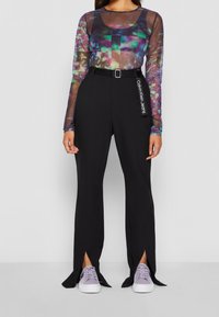 Doorzichtige top met lange mouwen en een abstract patroon in meerdere kleuren, gecombineerd met zwarte, taillehoge broek met zijsplitsingen en een kleine riem.