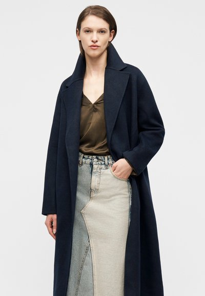 Γυναίκα με σκούρο μπλε σκούφο παλτό πάνω από μια καφέ μεταξωτή μπλούζα και μια ψηλόμεση δίχρωμη φούστα από τζιν, με το ένα χέρι στην τσέπη.