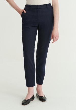 Femme portant un pantalon slim bleu marine longueur cheville et des ballerines noires, debout avec une main dans la poche, sur un fond uni.