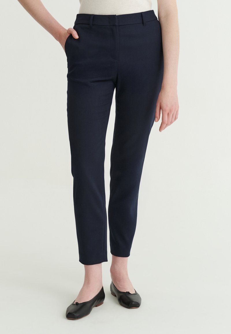 Femme portant un pantalon slim bleu marine longueur cheville et des ballerines noires, debout avec une main dans la poche, sur un fond uni.