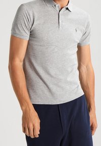 Man wearing a light gray short-sleeve polo shirt with buttons and navy blue pants, standing against a plain background.