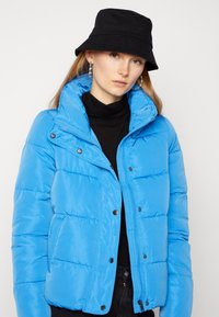 Veste rembourrée bleu clair avec un motif matelassé, col montant et boutons-pression. Portée sur un col roulé noir, accessoirisée avec un chapeau seau noir.
