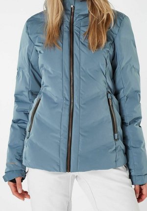 Vrouw draagt een lichtblauwe doorgestikte jas met zwarte ritssluiting en zijritszakken, gecombineerd met een witte broek.