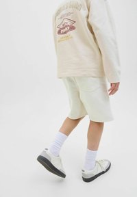 Sudadera beige con texto gráfico, pantalones cortos crema, calcetines blancos y zapatillas beige con suelas negras. Enfoque en un diseño simple y casual.