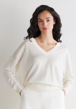 Kvinde med mørkt krøllet hår iført en løs hvid V-hals sweater og hvide bukser med hænderne i lommerne mod en ensartet lys baggrund.