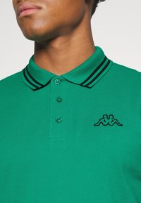 Zielona koszulka polo o teksturowanym wykończeniu, z kołnierzykiem i guzikami w czarne paski. Zawiera małe czarne logo na lewej piersi.