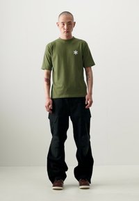Oliven grøn bomulds t-shirt med et hvidt stjernesymbol på brystet, parret med løstsiddende sorte cargobukser og brune sko.