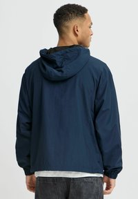 Veste à capuche bleu marine en matériau léger, avec des poignets élastiques et une coupe décontractée. La vue de dos montre une texture unie sans motifs.