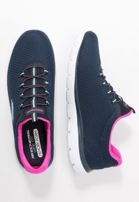 Námořnicky modré tenisky Skechers s širokým střihem a stélkou z paměťové pěny, s růžovým vnitřním lemem, tyrkysovými dekorativními pruhy a bílými podrážkami, zobrazené z horního a bočního úhlu.