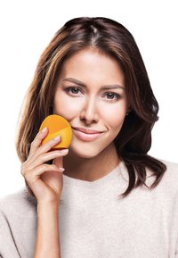 Orange silikon ansiktsrengöringsapparat med texturerad yta, hålls av en kvinna med mellanmörkt brunt hår, klädd i en ljusbeige tröja.