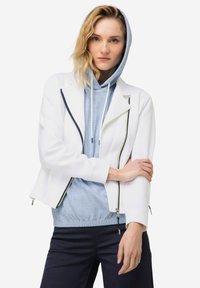 Chaqueta blanca corta con detalles en negro en el cierre, superpuesta sobre una sudadera a rayas azul claro con cordón y dobladillo acanalado.