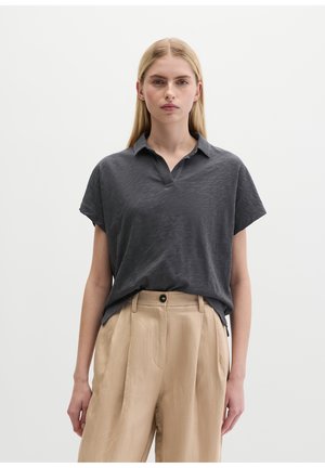Femme portant un polo gris foncé à manches courtes, rentré dans un pantalon beige plissé taille haute, sur un fond uni.