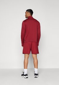Sudadera atlética burdeos y pantalones cortos; la sudadera tiene un cuello alto y una textura suave; los pantalones cortos tienen un corte holgado y aberturas en los laterales.