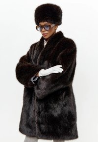 Žena v tmavě hnědém kožichu a odpovídající kožichové čepici, bílých rukavicích, modře tónovaných slunečních brýlích a světle šedých kalhotách na bílém pozadí.